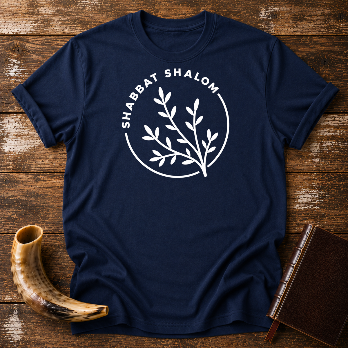 Navy t-shirt with a white SHABBAT SHALOM circle around a leafy olive branch, laid flat with a shofar and a leather book