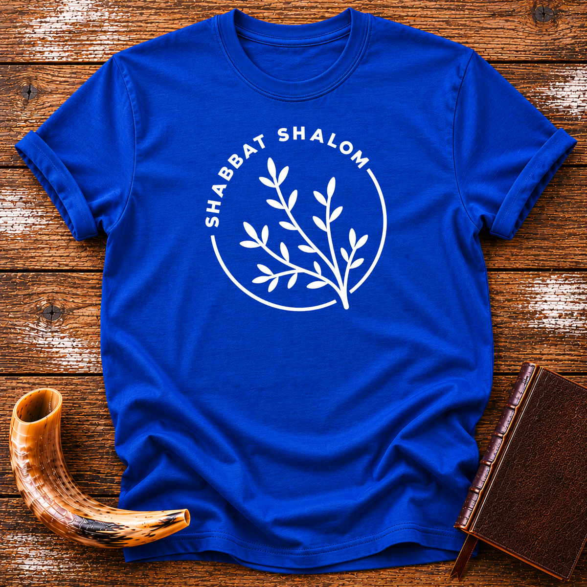 Royal blue t-shirt with a white SHABBAT SHALOM circle around a leafy olive branch, laid flat with a shofar and a leather book