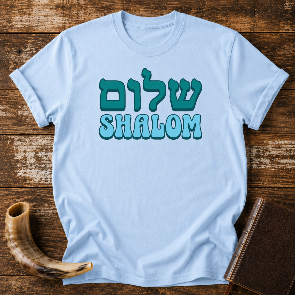 Light blue t-shirt with teal Hebrew lettering above SHALOM in blue bubble letters, laid flat with a shofar and a leather book