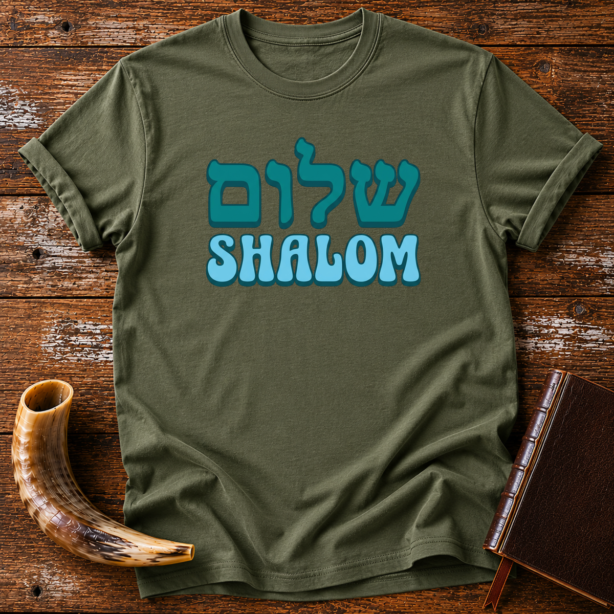Military green t-shirt with teal Hebrew lettering above SHALOM in blue bubble letters, laid flat with a shofar and a leather book