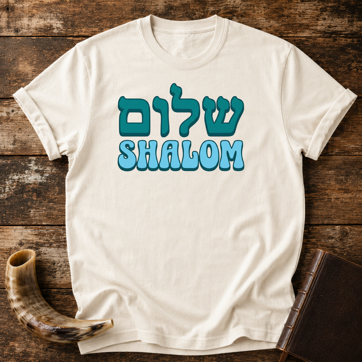 Natural cream t-shirt with teal Hebrew lettering above SHALOM in blue bubble letters, laid flat with a shofar and a leather book