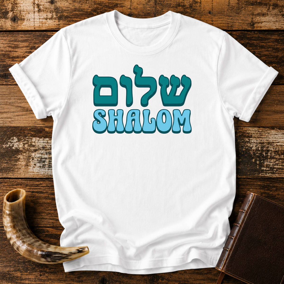 White t-shirt with teal Hebrew lettering above SHALOM in blue bubble letters, laid flat with a shofar and a leather book