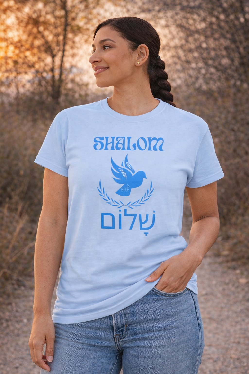 Light blue Shalom dove t-shirt with a blue dove, laurel and Hebrew lettering, worn by a woman outdoors