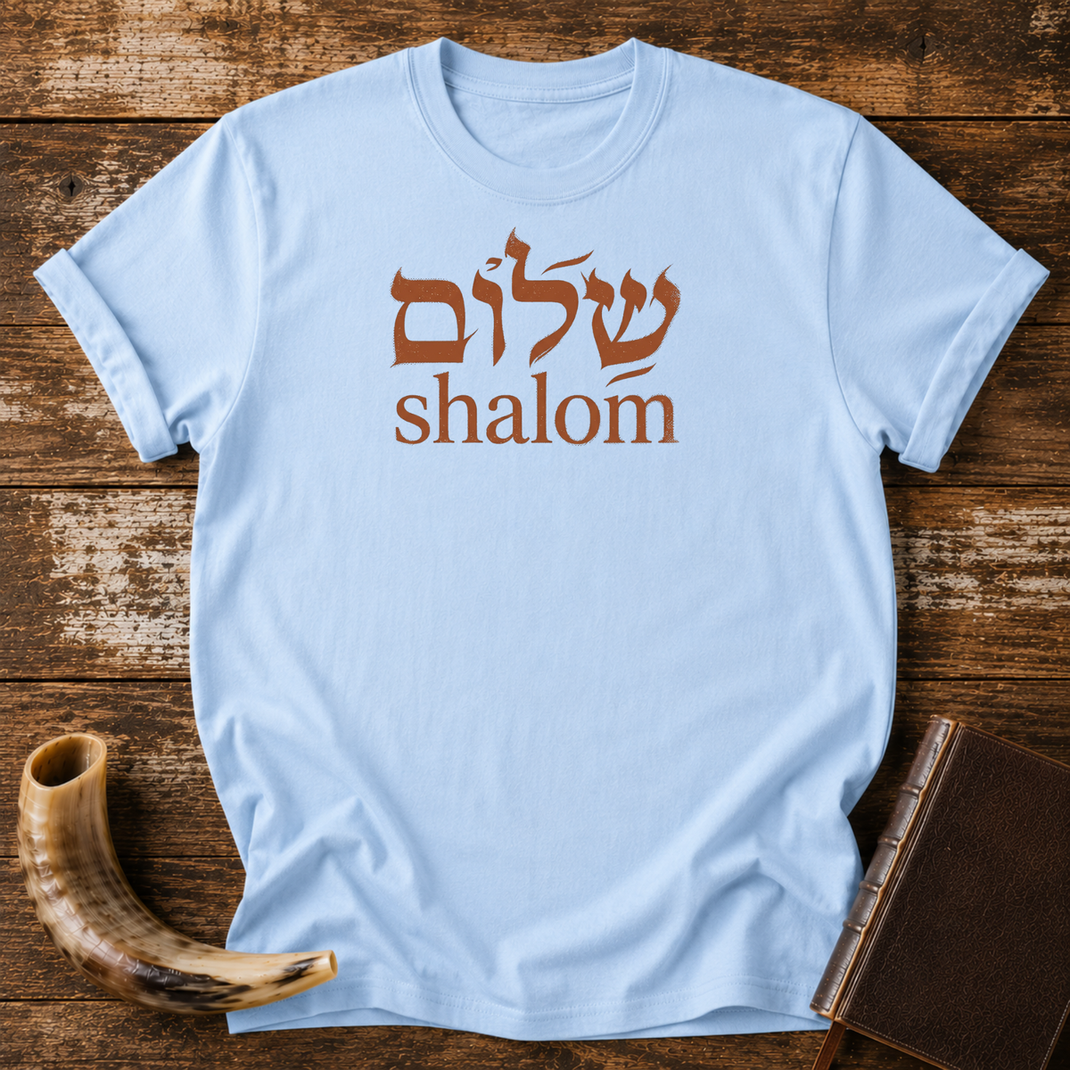 Light blue t-shirt with rust-colored Hebrew lettering above the word shalom, laid flat with a shofar and a leather book