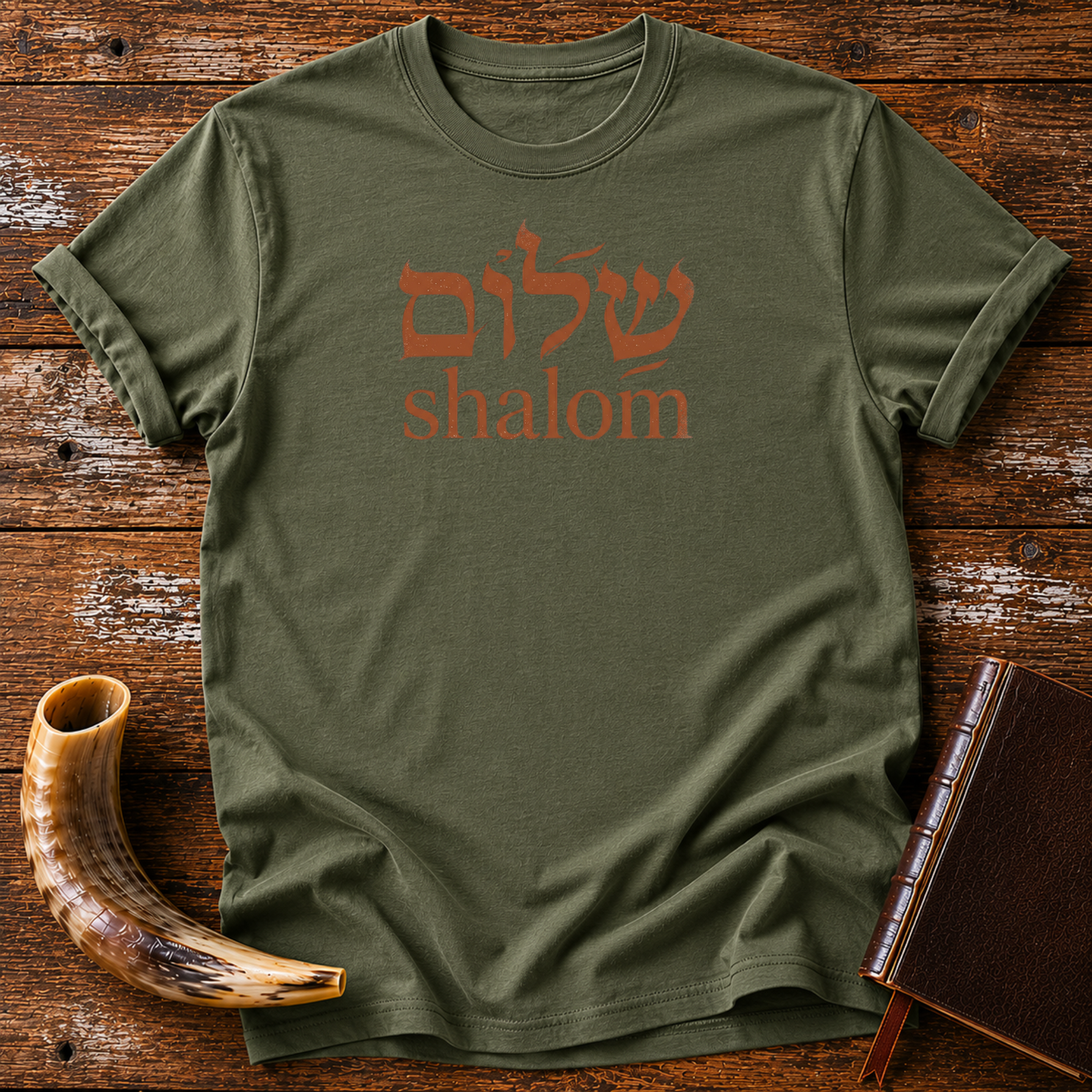 Military green t-shirt with rust-colored Hebrew lettering above the word shalom, laid flat with a shofar and a leather book