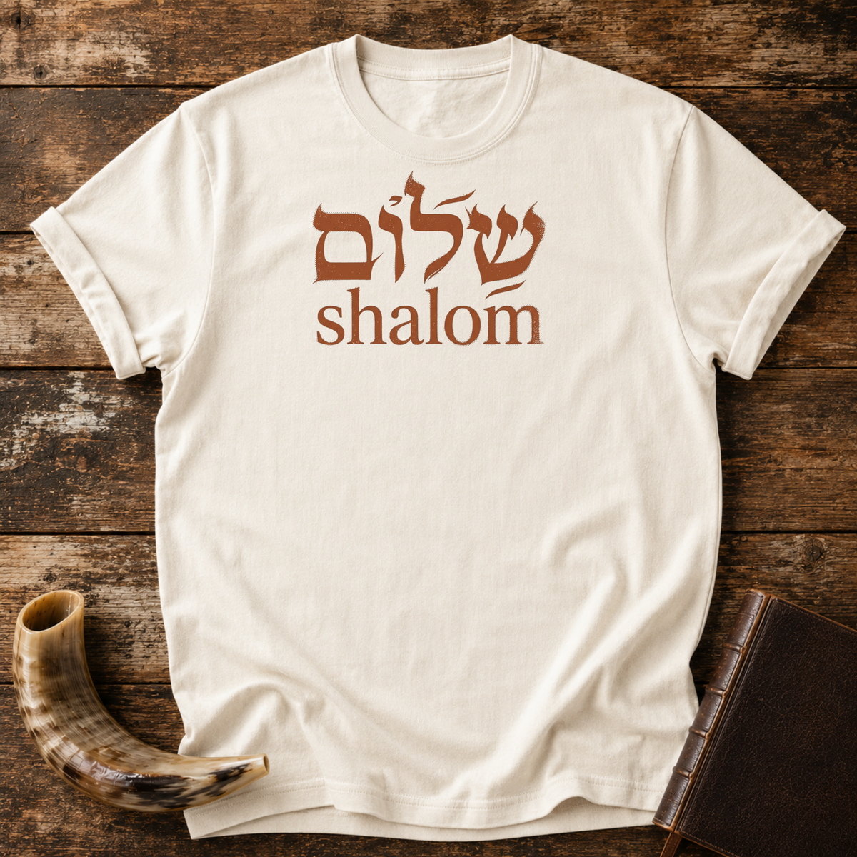Natural cream t-shirt with rust-colored Hebrew lettering above the word shalom, laid flat with a shofar and a leather book