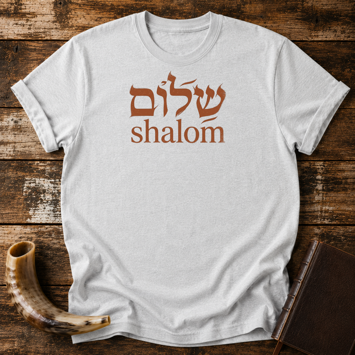 Sport grey t-shirt with rust-colored Hebrew lettering above the word shalom, laid flat with a shofar and a leather book