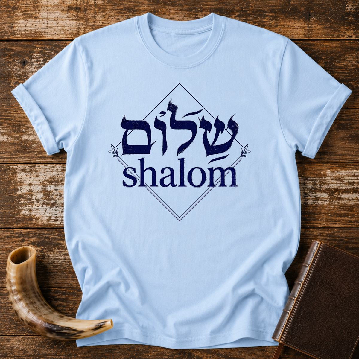 Light blue t-shirt with navy Hebrew lettering and the word shalom inside a diamond outline, laid flat with a shofar and a leather book