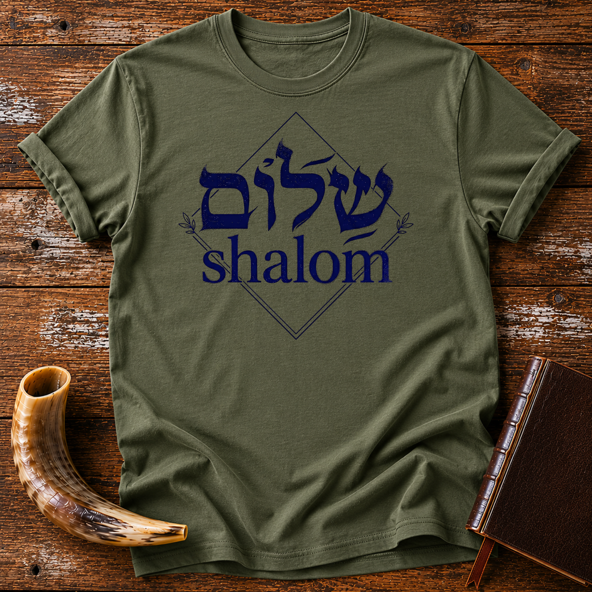 Military green t-shirt with navy Hebrew lettering and the word shalom inside a diamond outline, laid flat with a shofar and a leather book
