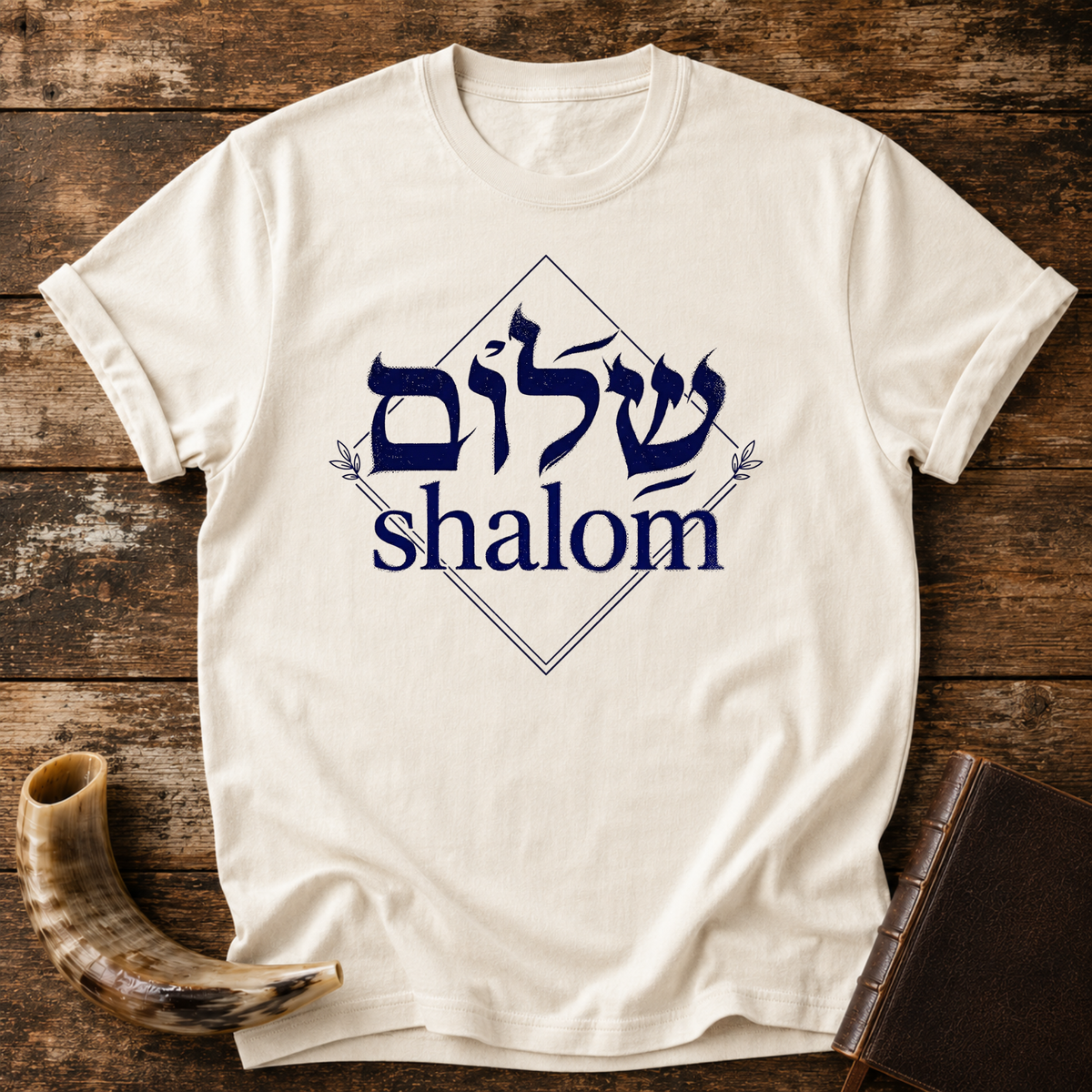 Natural cream t-shirt with navy Hebrew lettering and the word shalom inside a diamond outline, laid flat with a shofar and a leather book