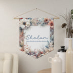Shalom wall pennant framed in watercolor flowers with Peace I leave with you, my peace I give unto you, hanging on a cream wall beside vases