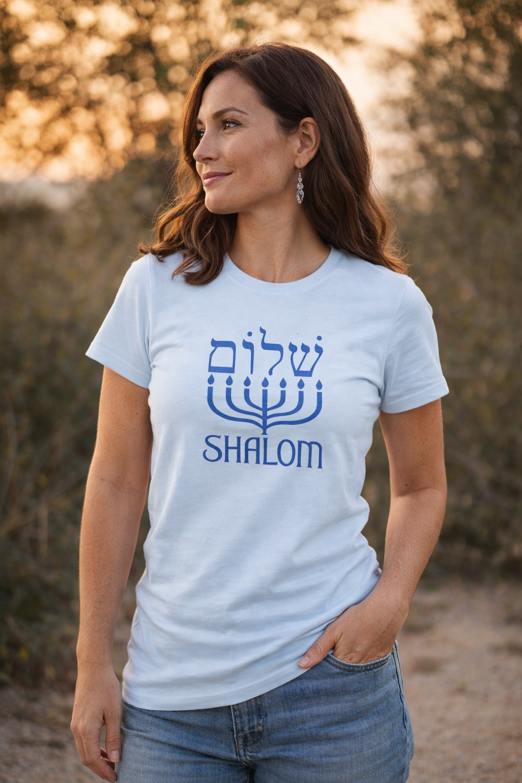 Light blue Shalom t-shirt with blue Hebrew lettering above a menorah, worn by a woman on an outdoor path at sunset