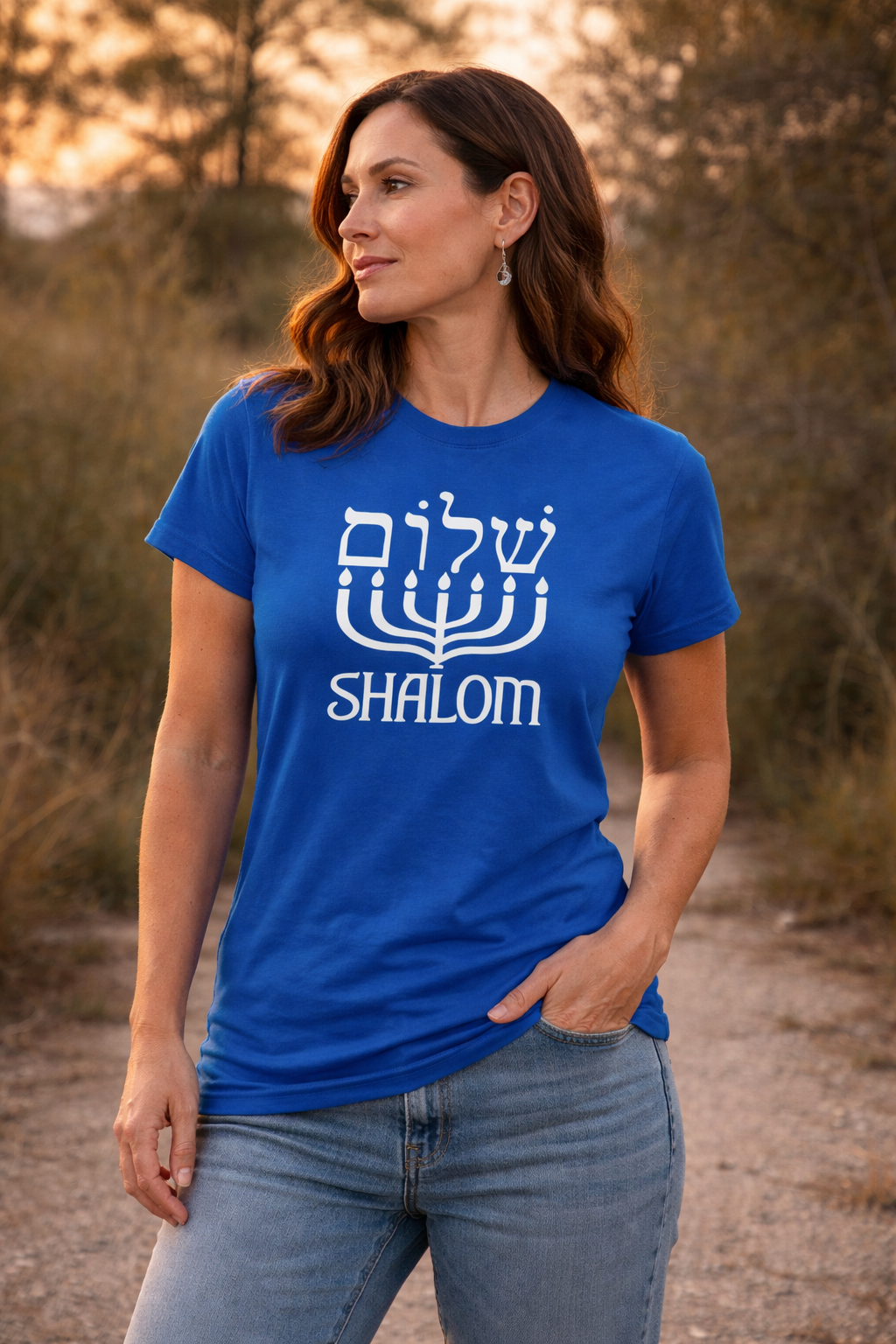 Royal blue Shalom t-shirt with white Hebrew lettering above a menorah, worn by a woman on a path at sunset