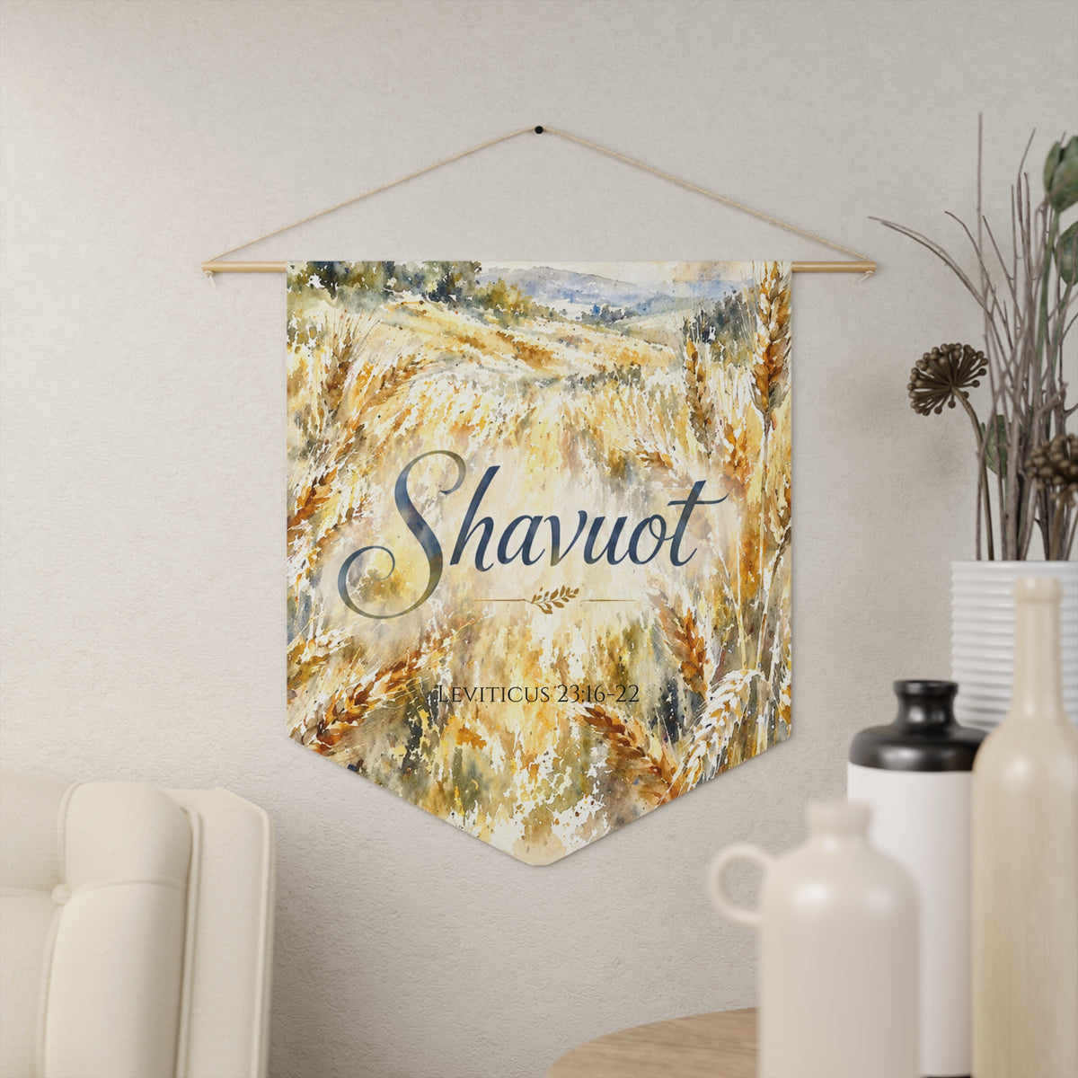 Watercolor wheat field Shavuot wall pennant with blue script lettering, Leviticus 23:16-22, hanging on a cream wall beside vases