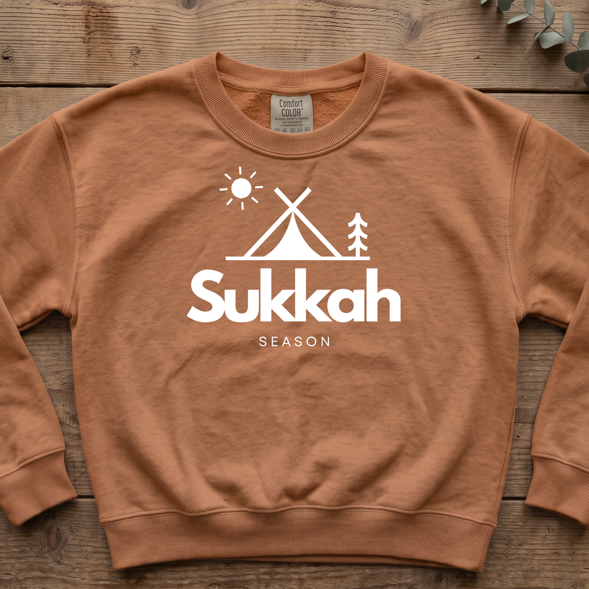 Yam orange Sukkah Season sweatshirt with a simple tent, sun and pine tree graphic, laid flat on wood boards