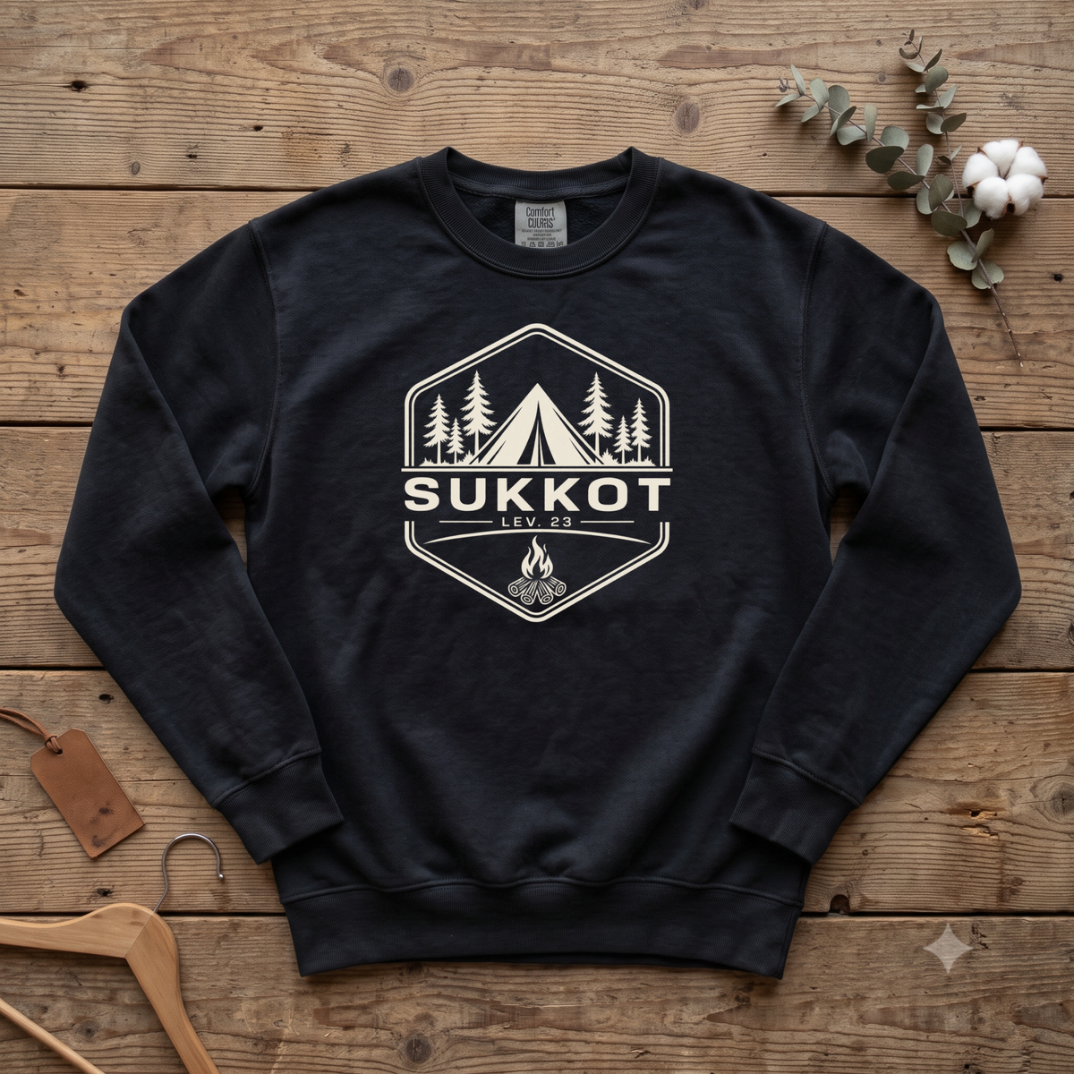 Black SUKKOT Lev. 23 camp badge sweatshirt with a tent, pines and campfire, laid flat on a wooden table