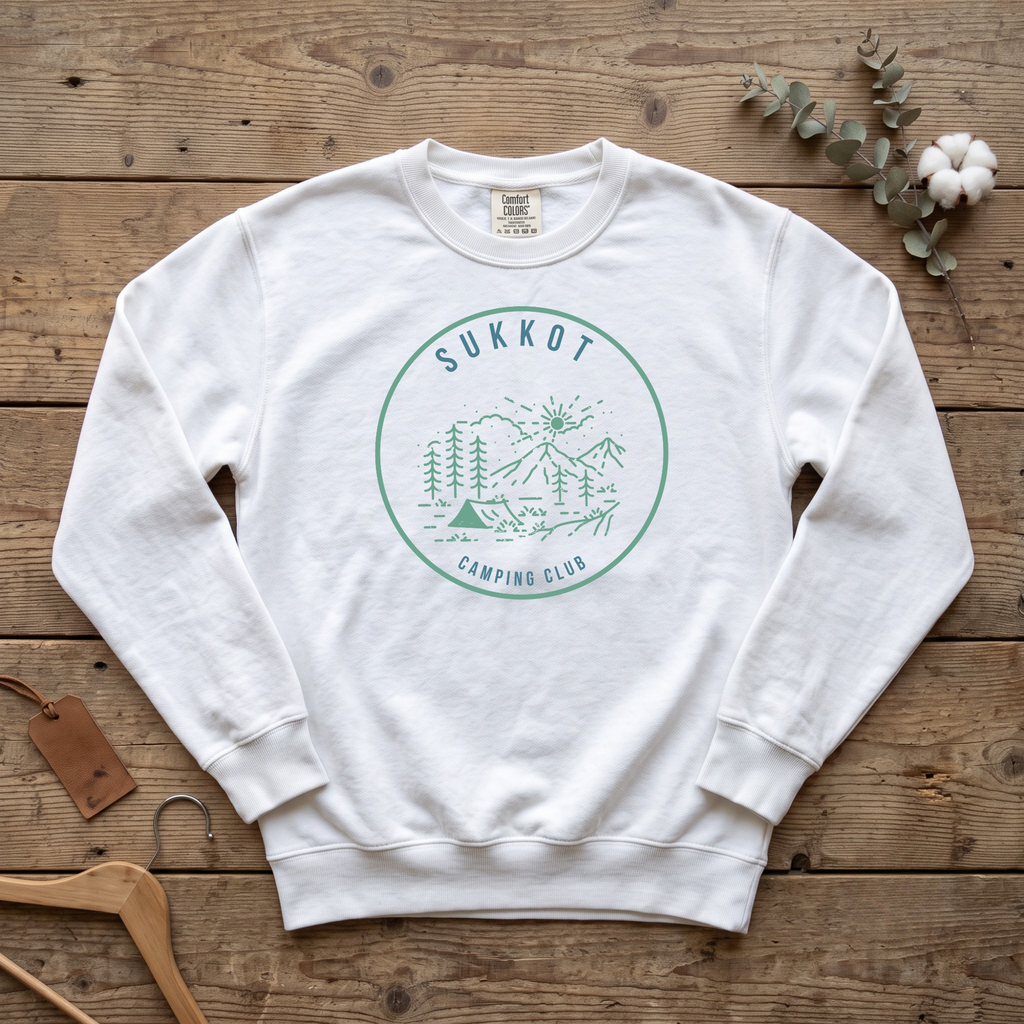 White SUKKOT Camping Club sweatshirt with a green circle emblem of a tent, pines and mountains, laid flat on a wooden table
