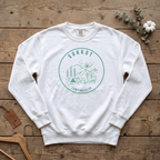 White SUKKOT Camping Club sweatshirt with a green circle emblem of a tent, pines and mountains, laid flat on a wooden table