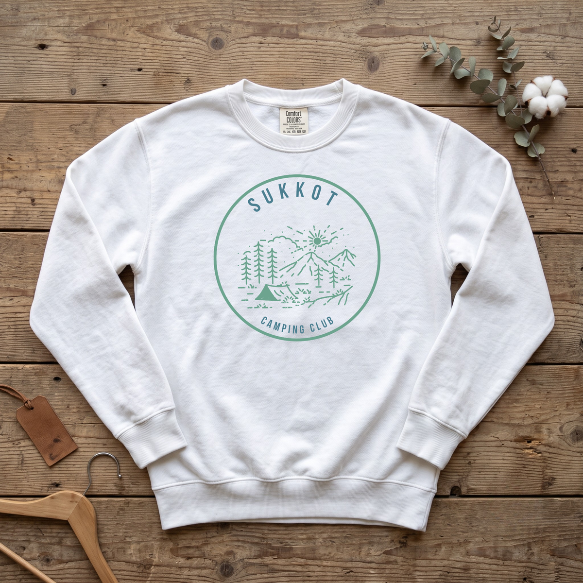 White SUKKOT Camping Club sweatshirt with a green circle emblem of a tent, pines and mountains, laid flat on a wooden table
