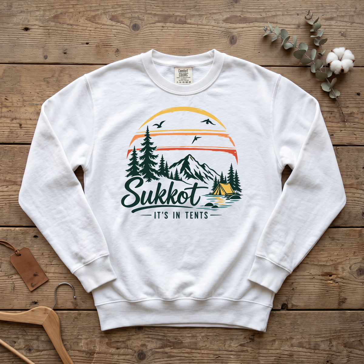 White Sukkot It's In Tents sweatshirt with a sunset, mountains, pines and a tent, laid flat on a wooden table
