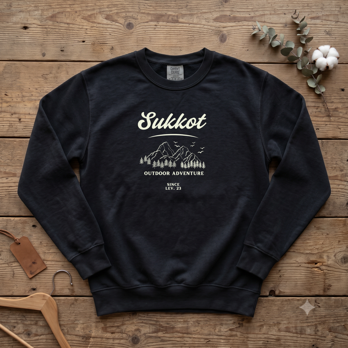 Black Sukkot Outdoor Adventure Since Lev. 23 sweatshirt with a mountain and pines sketch, laid flat on a wooden table