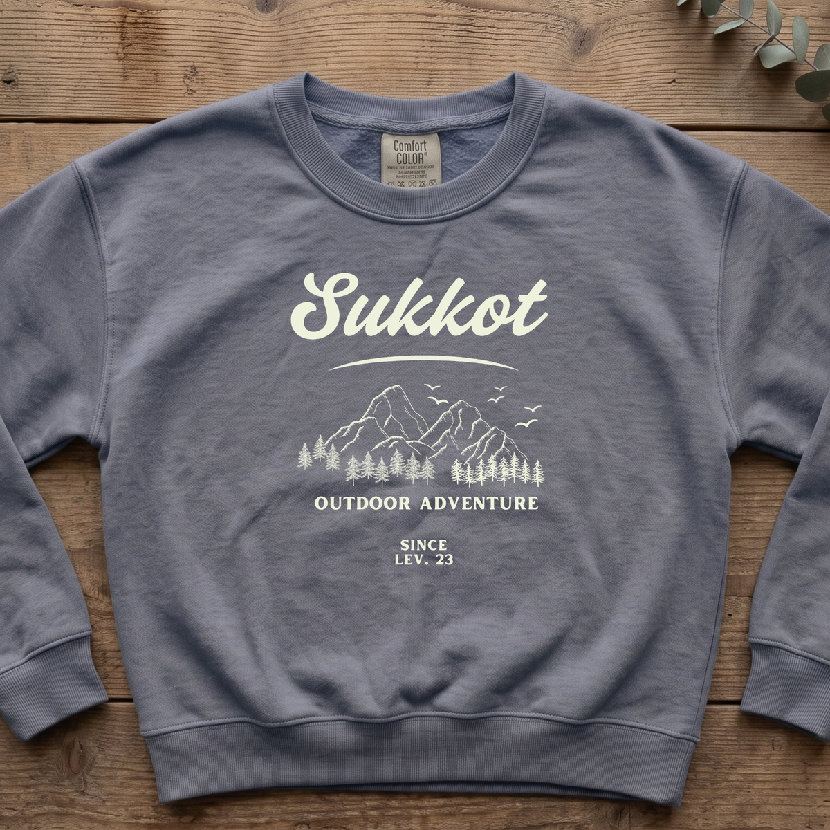 Blue jean Sukkot Outdoor Adventure Since Lev. 23 sweatshirt with a mountain and pines sketch, laid flat on a wooden table