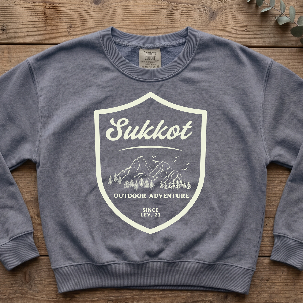 Blue jean Sukkot Outdoor Adventure Since Lev. 23 sweatshirt with a mountain shield badge, laid flat on wood boards