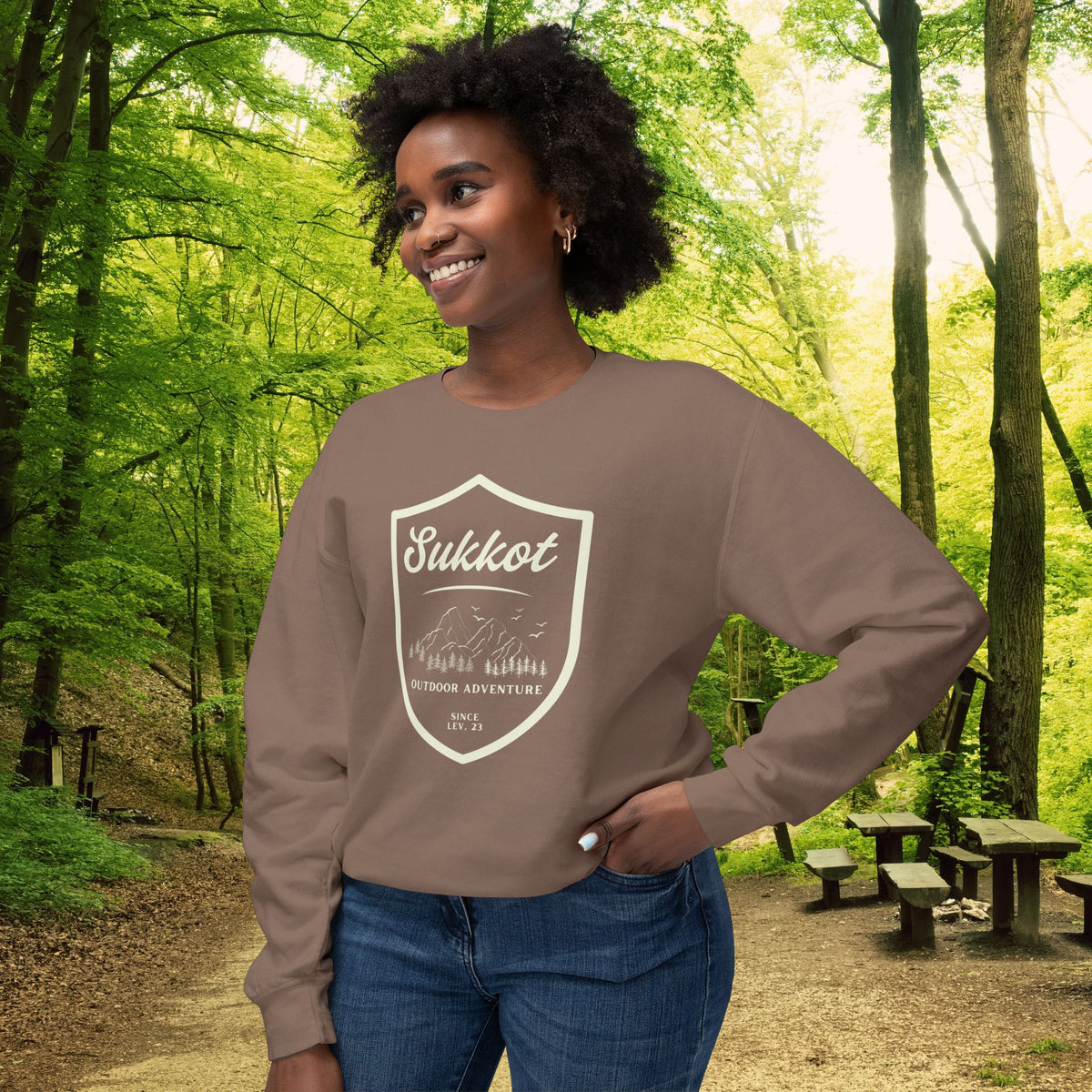 Espresso brown Sukkot Outdoor Adventure Since Lev. 23 sweatshirt with a mountain shield badge, worn by a smiling woman in a forest