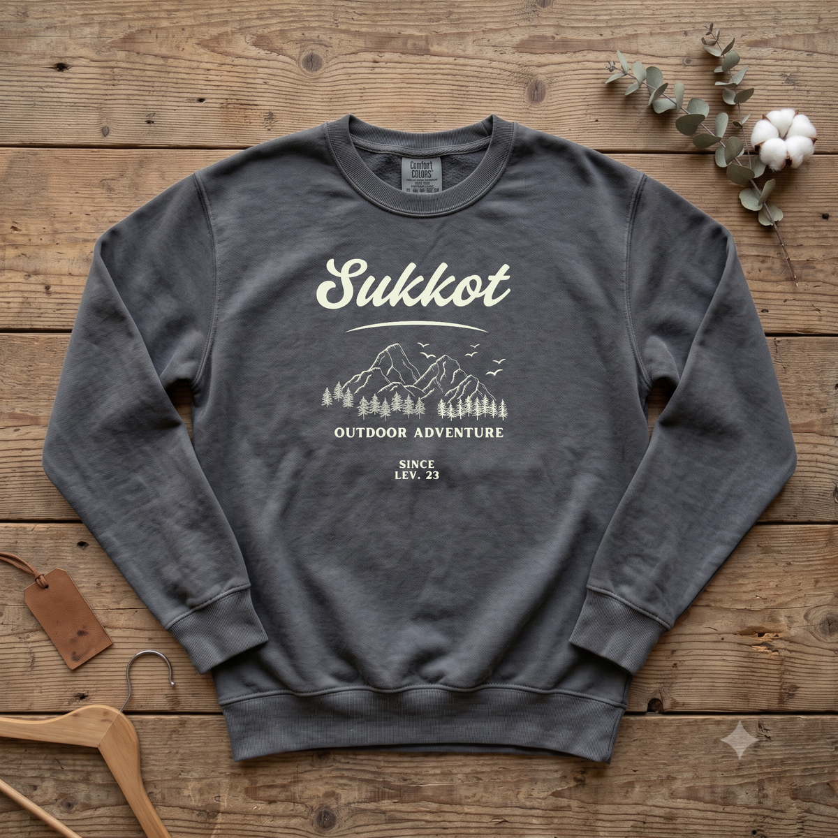 Pepper Sukkot Outdoor Adventure Since Lev. 23 sweatshirt with a mountain and pines sketch, laid flat on a wooden table