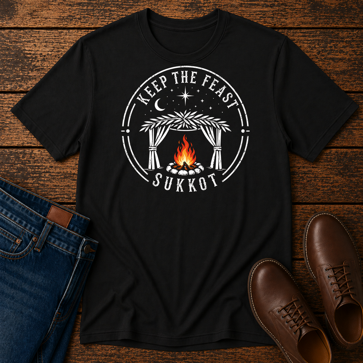 Black t-shirt with a white KEEP THE FEAST SUKKOT sukkah badge with an orange campfire, laid flat with jeans and brown leather boots
