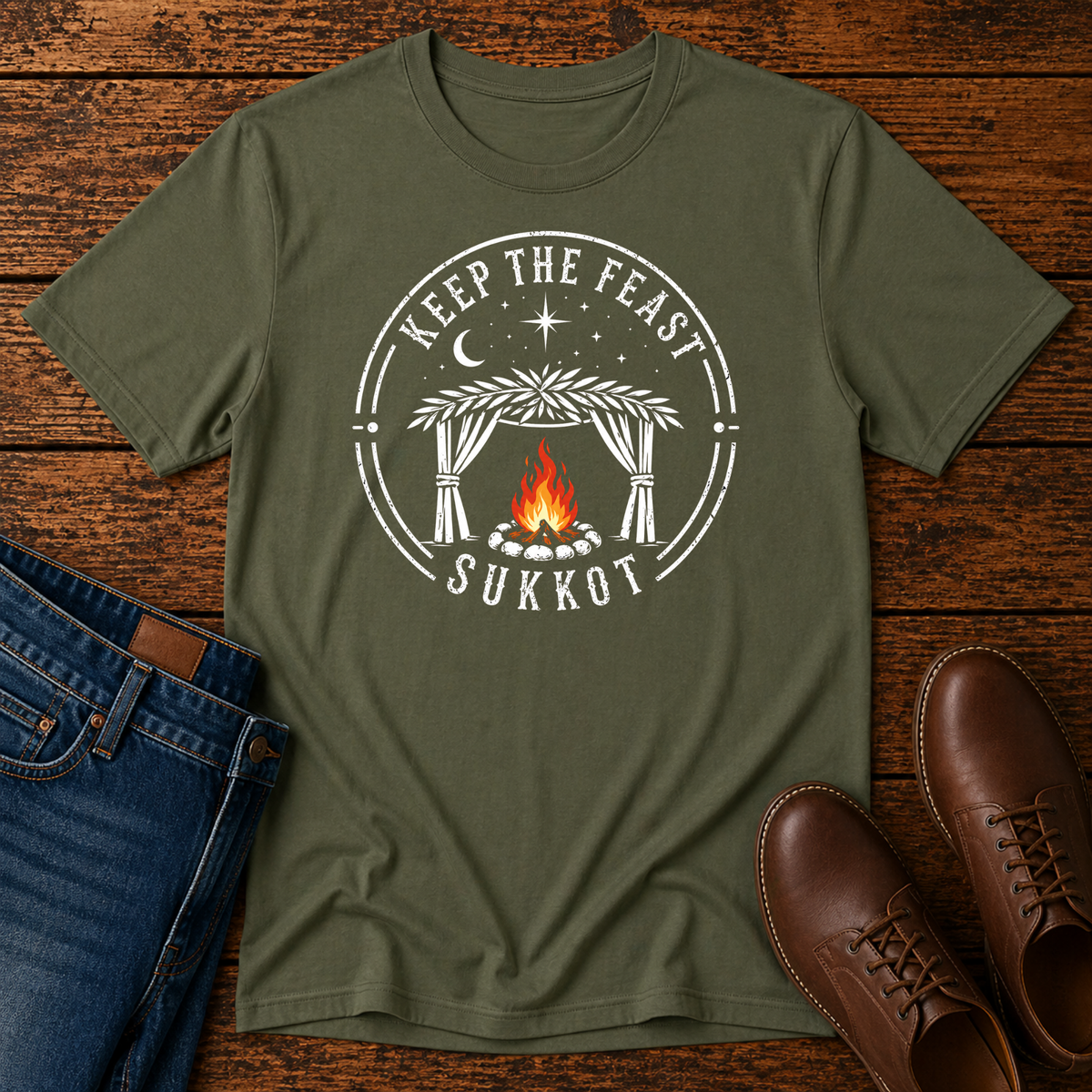 Military green t-shirt with a white KEEP THE FEAST SUKKOT sukkah badge with an orange campfire, laid flat with jeans and brown leather boots