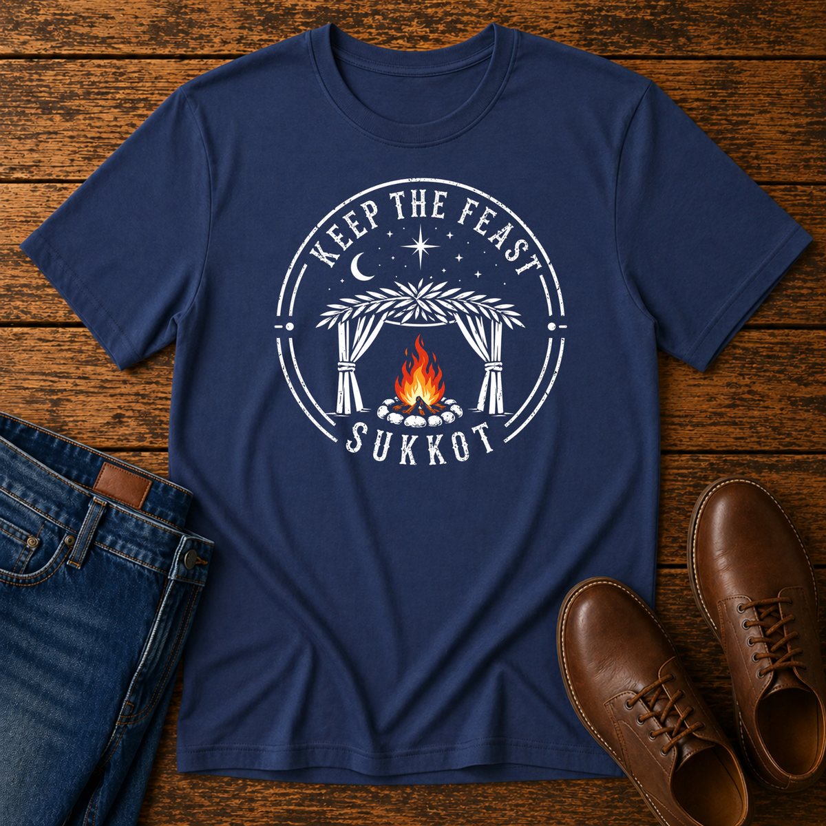 Navy t-shirt with a white KEEP THE FEAST SUKKOT sukkah badge with an orange campfire, laid flat with jeans and brown leather boots