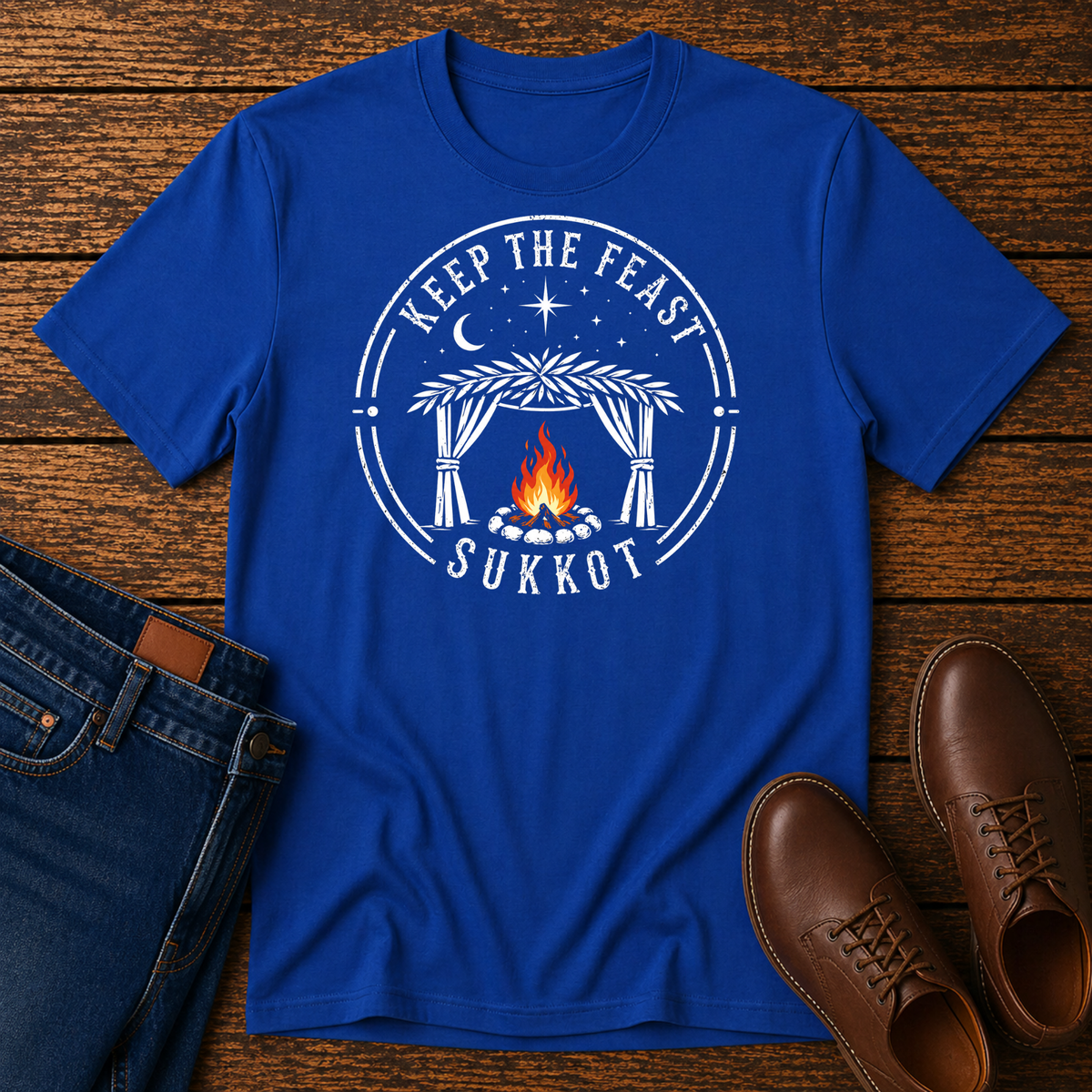 Royal blue t-shirt with a white KEEP THE FEAST SUKKOT sukkah badge with an orange campfire, laid flat with jeans and brown leather boots