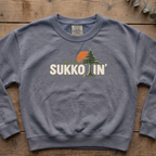 Blue jean Sukkotin' sweatshirt with a pine tree and orange sun in the lettering, laid flat on a wooden table