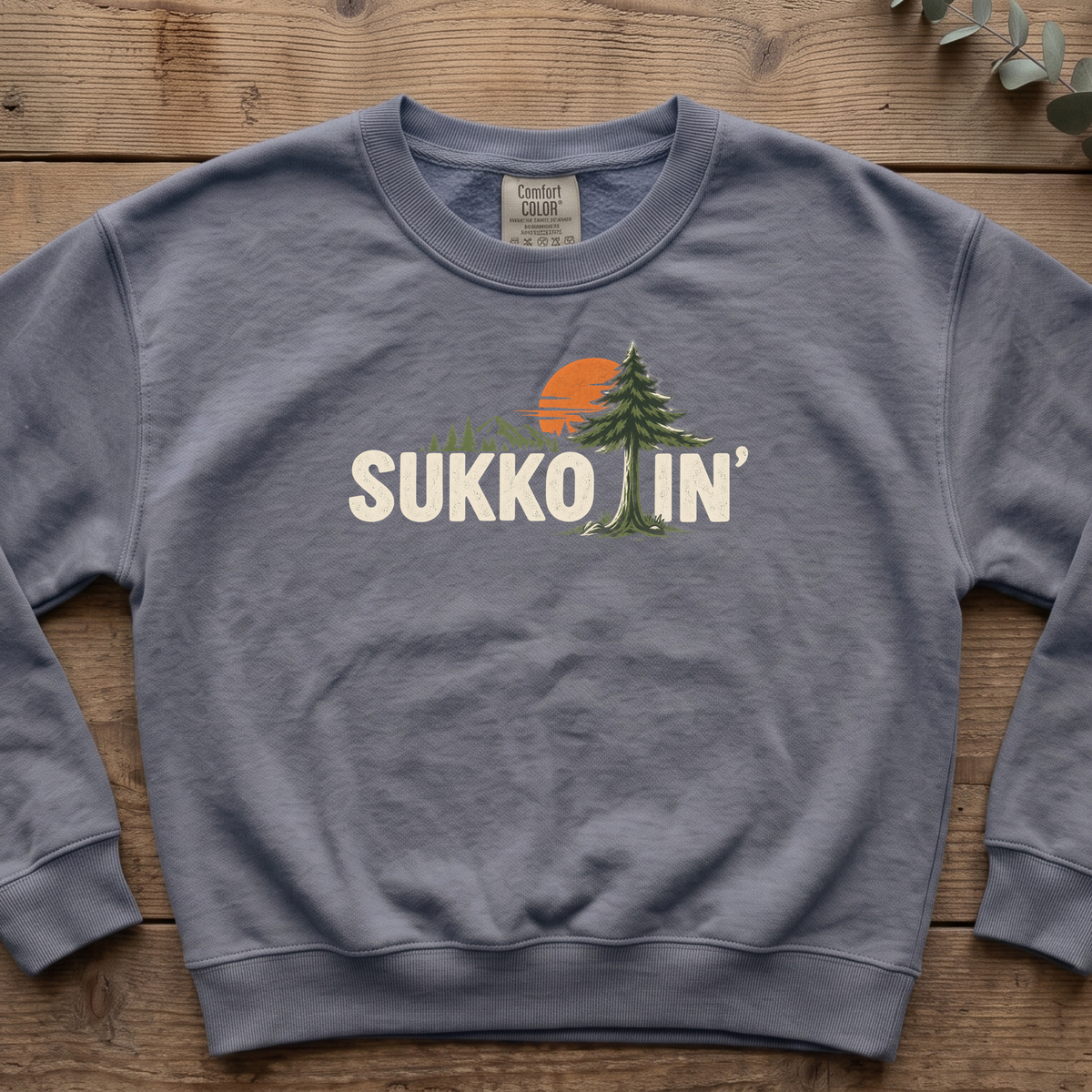 Blue jean Sukkotin' sweatshirt with a pine tree and orange sun in the lettering, laid flat on a wooden table