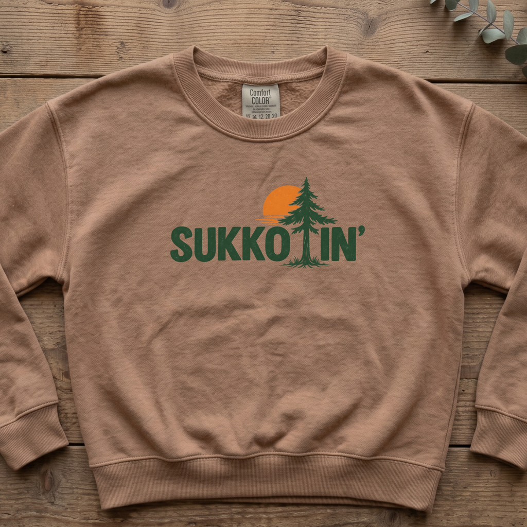 Espresso Sukkotin' sweatshirt with a pine tree and orange sun in the lettering, laid flat on a wooden table
