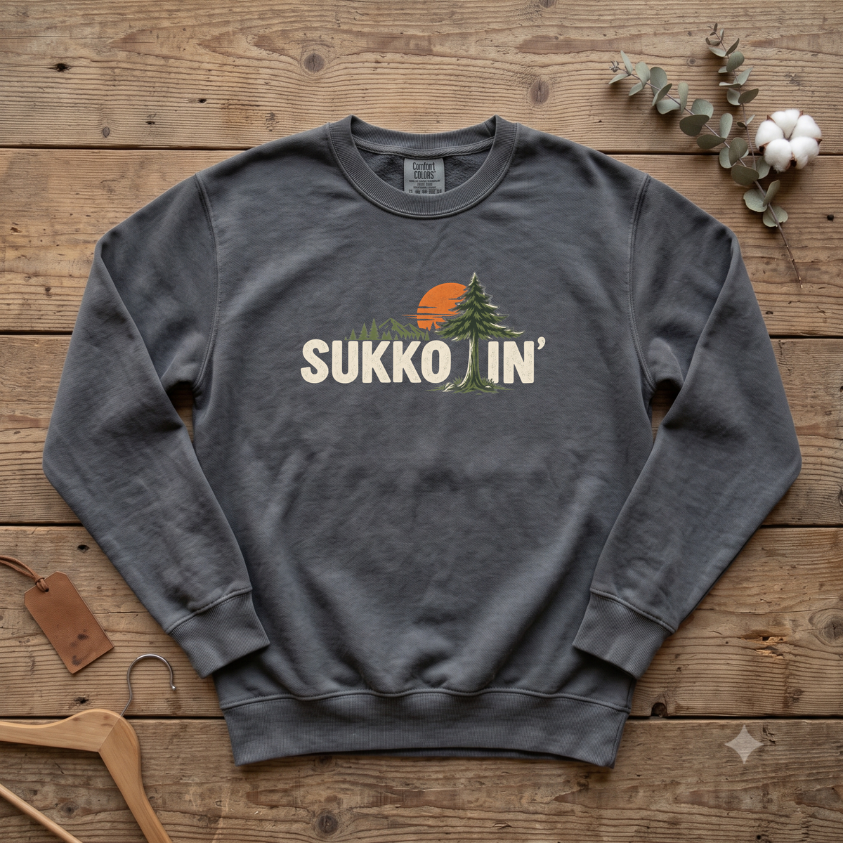 Pepper Sukkotin' sweatshirt with a pine tree and orange sun in the lettering, laid flat on a wooden table