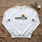 White Sukkotin' sweatshirt with a pine tree and orange sun in the lettering, laid flat on a wooden table