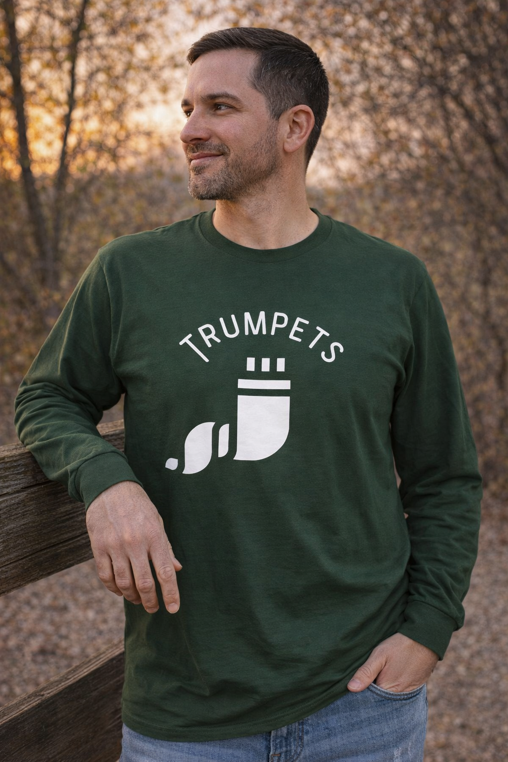 Forest green Trumpets long sleeve tee with a white shofar graphic, worn by a man leaning on a fence in autumn