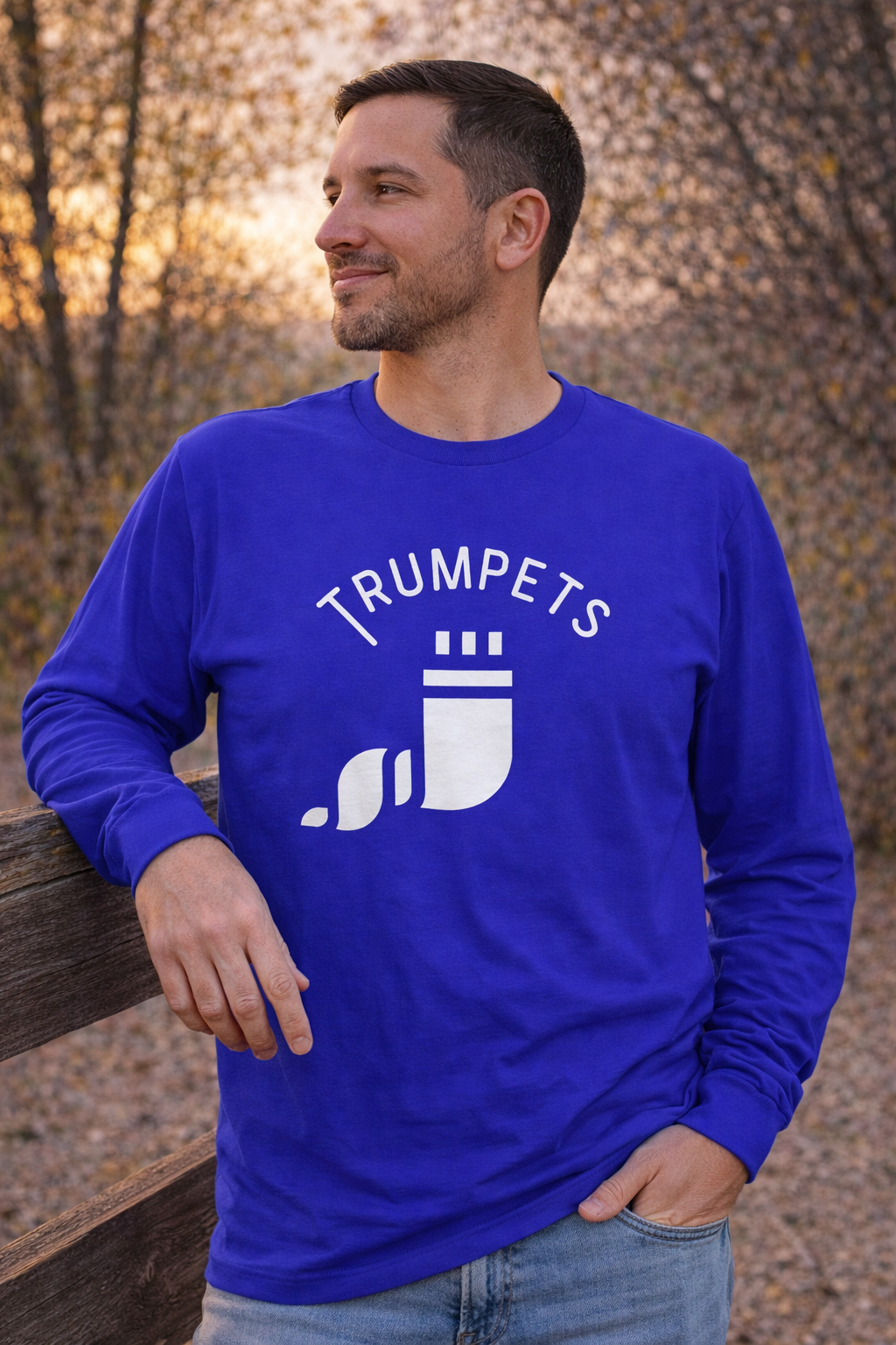 Royal blue Trumpets long sleeve tee with a white shofar graphic, worn by a man leaning on a fence in autumn