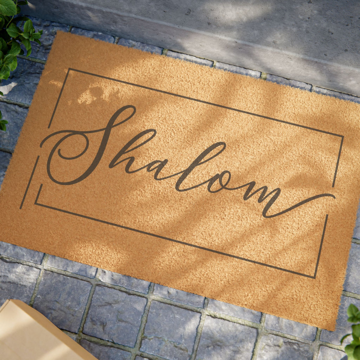 Natural coir doormat with "Shalom" in black script inside a thin frame, on a stone porch
