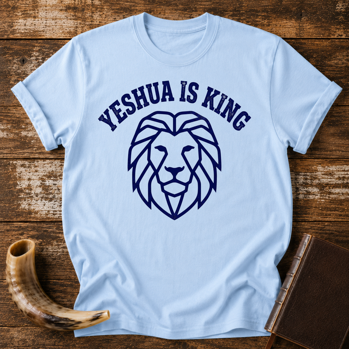 Light blue t-shirt with YESHUA IS KING arched over a navy line-art lion head, laid flat with a shofar and a leather book