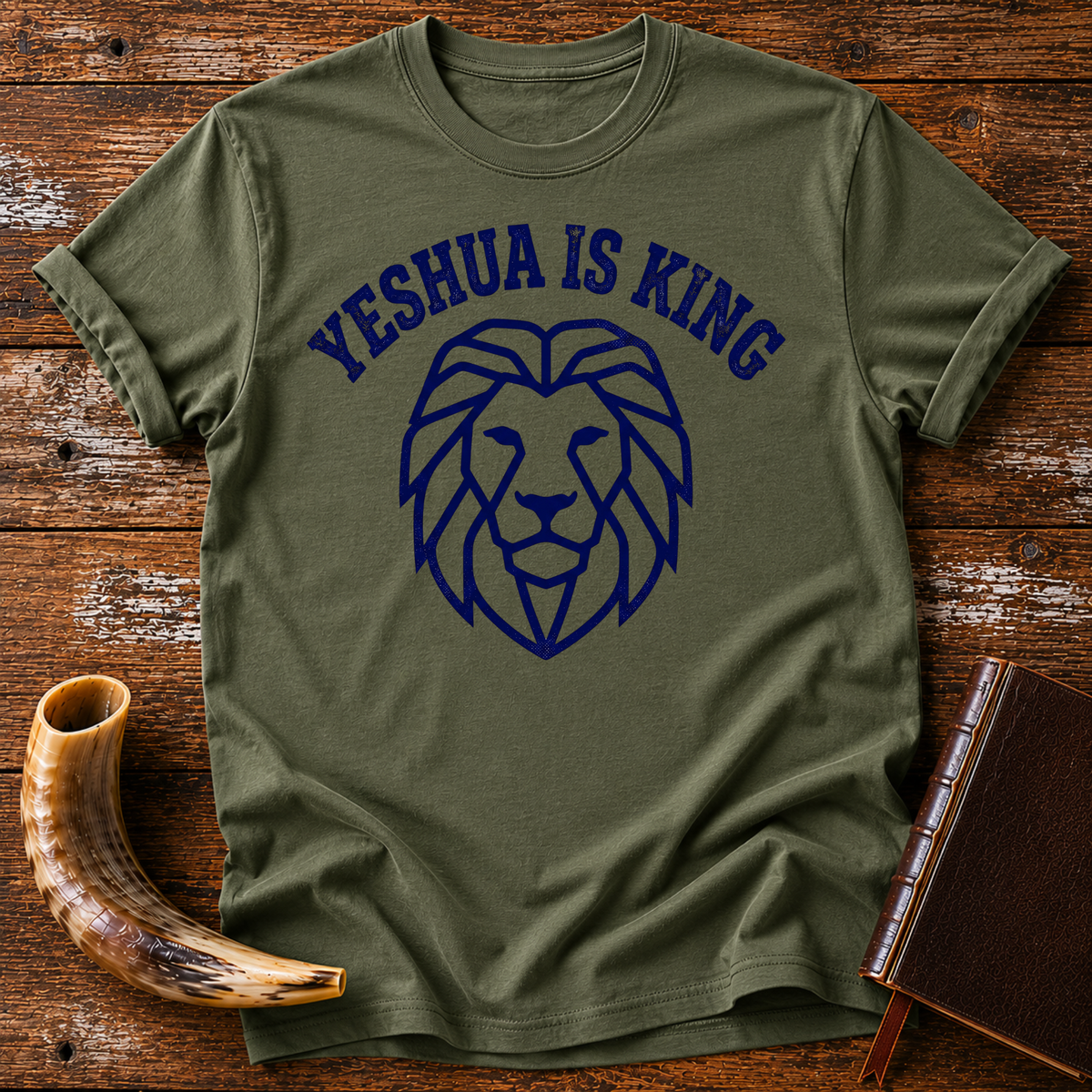 Military green t-shirt with YESHUA IS KING arched over a navy line-art lion head, laid flat with a shofar and a leather book