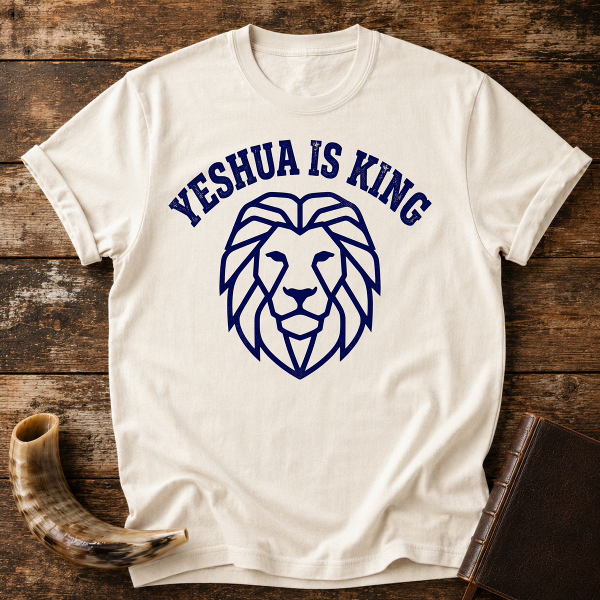 Natural cream t-shirt with YESHUA IS KING arched over a navy line-art lion head, laid flat with a shofar and a leather book