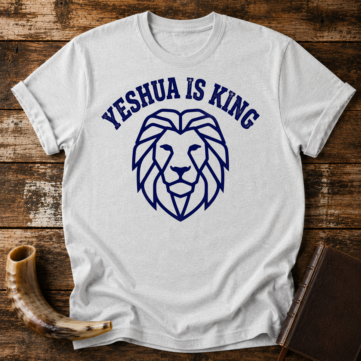 Sport grey t-shirt with YESHUA IS KING arched over a navy line-art lion head, laid flat with a shofar and a leather book