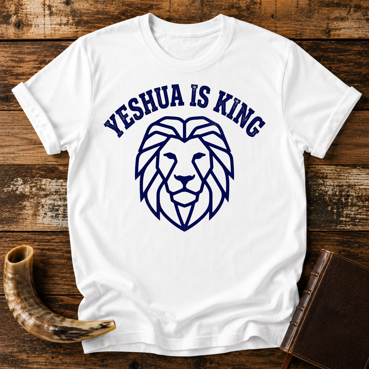 White t-shirt with YESHUA IS KING arched over a navy line-art lion head, laid flat with a shofar and a leather book
