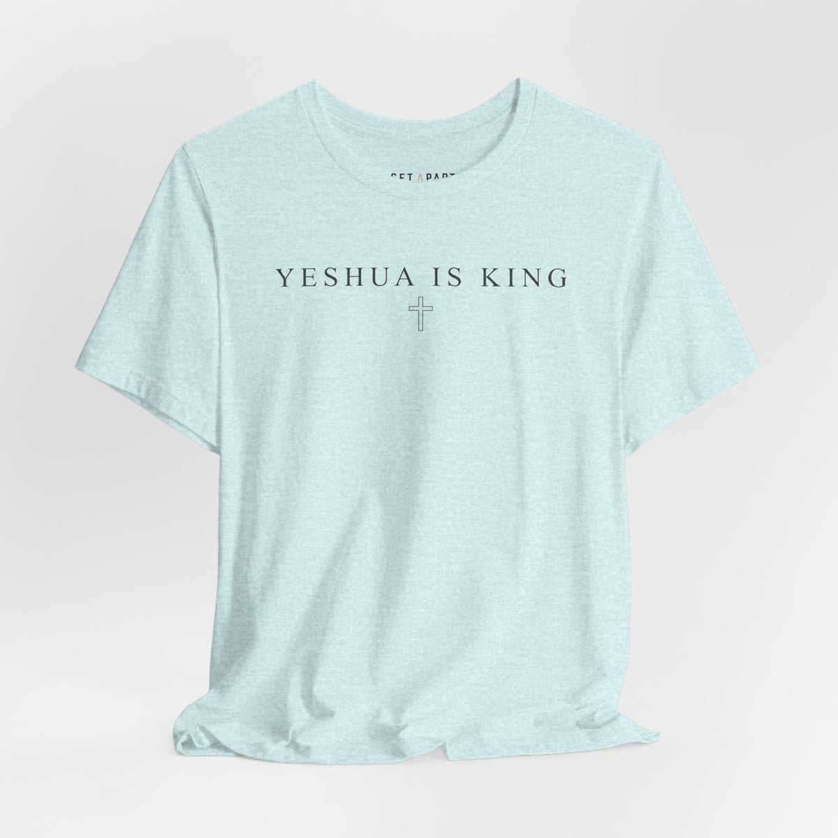 Heather ice blue t-shirt with small YESHUA IS KING text above a thin outlined cross, laid flat on a light gray background