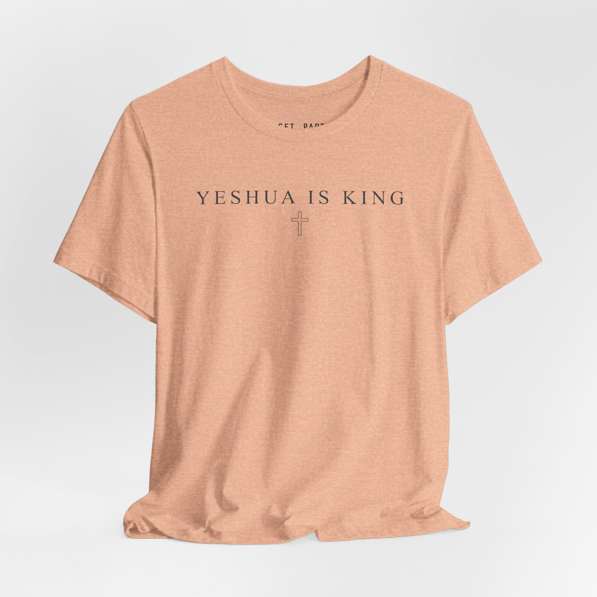 Heather peach t-shirt with small YESHUA IS KING text above a thin outlined cross, laid flat on a light gray background