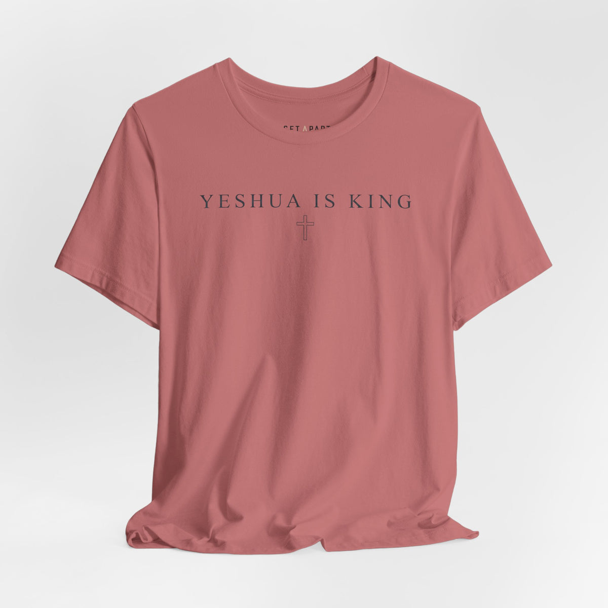 Mauve t-shirt with small YESHUA IS KING text above a thin outlined cross, shown front on a light gray background