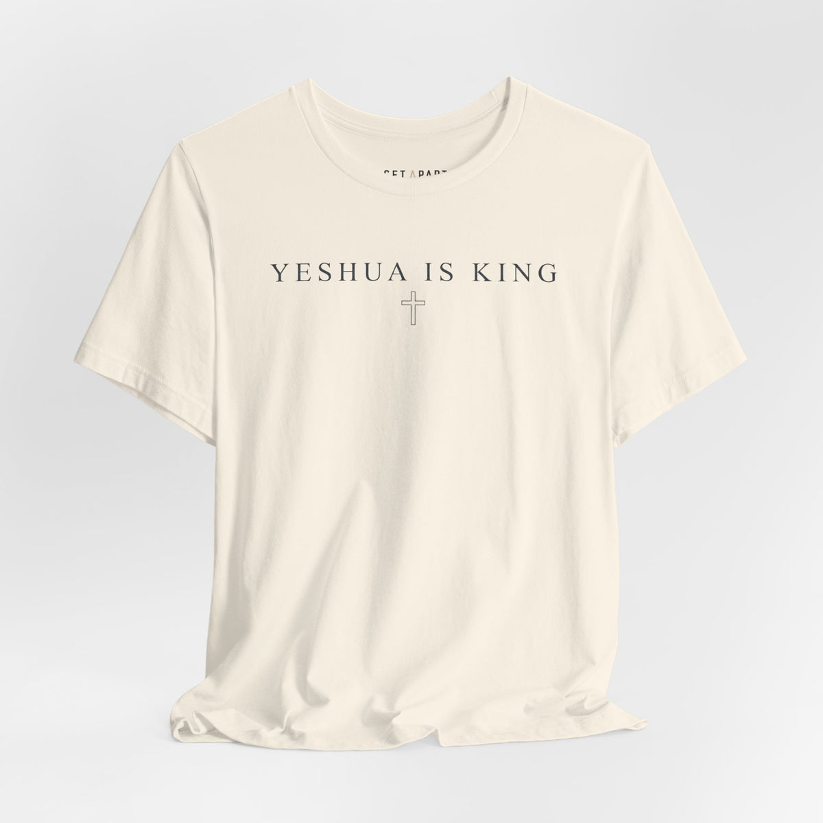 Natural cream t-shirt with small YESHUA IS KING text above a thin outlined cross, shown front on a light gray background